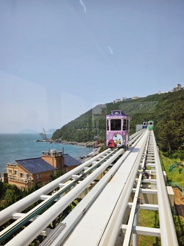 A colorful Sky Capsule traveling along the elevated coastal rail in Haeundae, Busan, with ocean and mountain scenery in the background.
