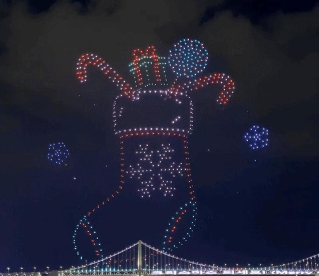 A Christmas stocking and ornaments created by illuminated drones above Gwangalli Beach, with the night view of Gwangan Bridge beneath.