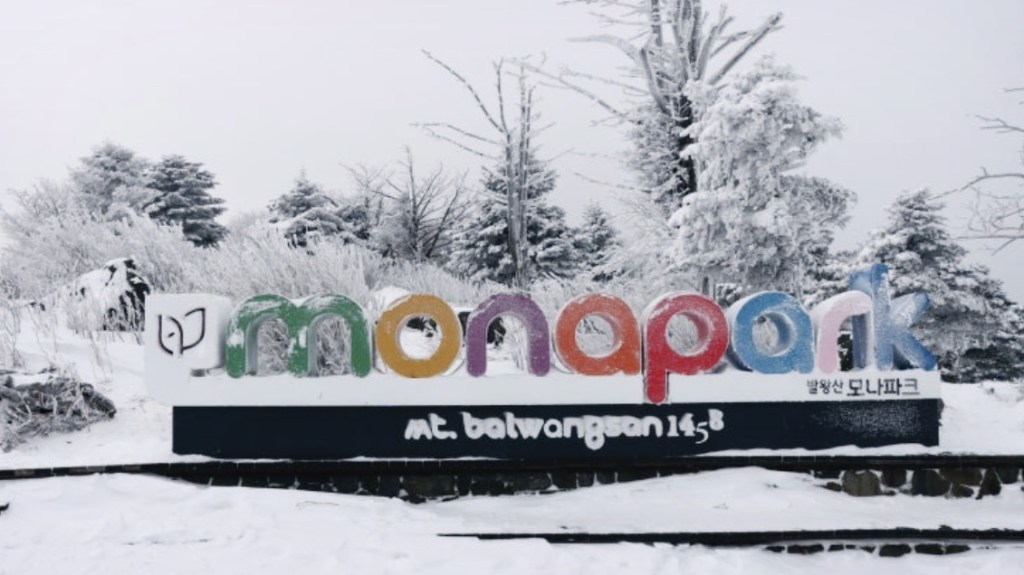 Snow-covered entrance sign of High1 Mona Park (Mt. Baekunsan), surrounded by winter trees in Gangwon-do, Korea
