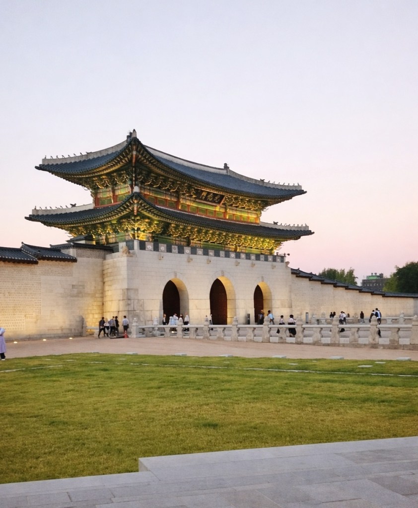 Gwanghwamun Gate in Seoul during early evening, showing traditional Korean architecture, a wide open lawn, and visitors walking along the historic palace walls.
