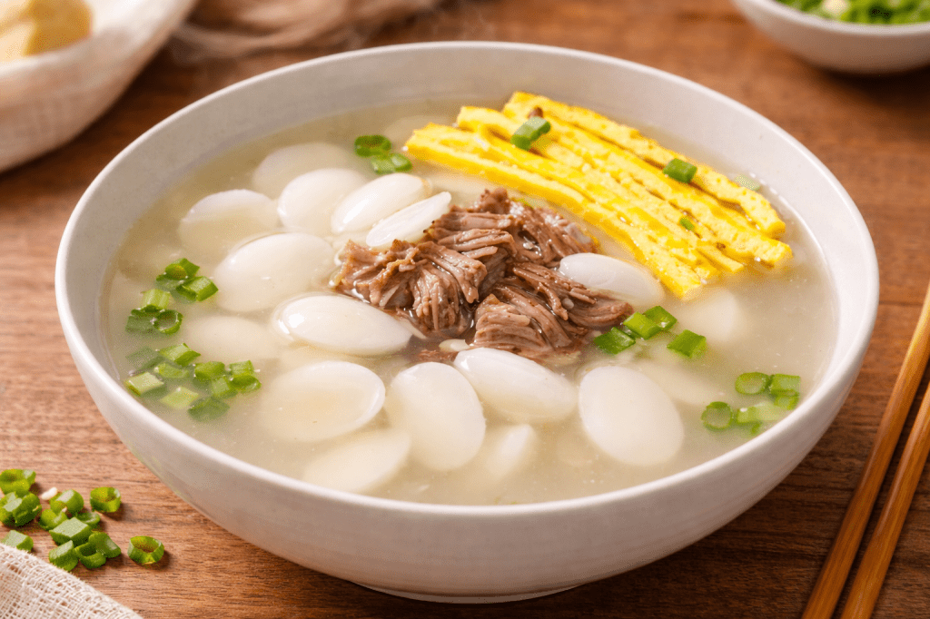 Korean tteokguk rice cake soup with clear white broth, sliced rice cakes, shredded beef, egg garnish, and seaweed in a ceramic bowl